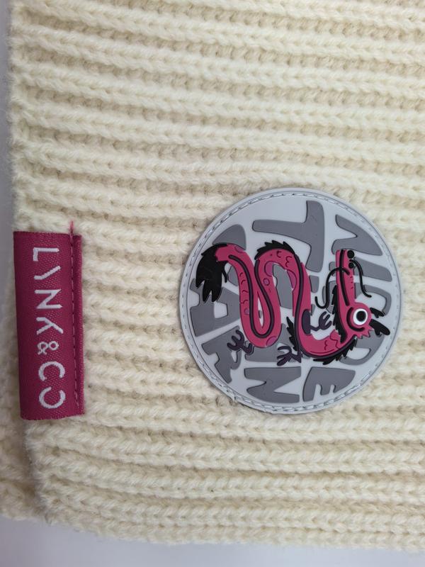 Crème grofgebreide muts met roze draak rubberen patch en roze merklabel op gestructureerde wol.
