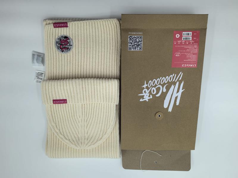 Crèmekleurige geribbelde gebreide muts en sjaal set met bordeauxrode logopatch naast geschenkverpakking.