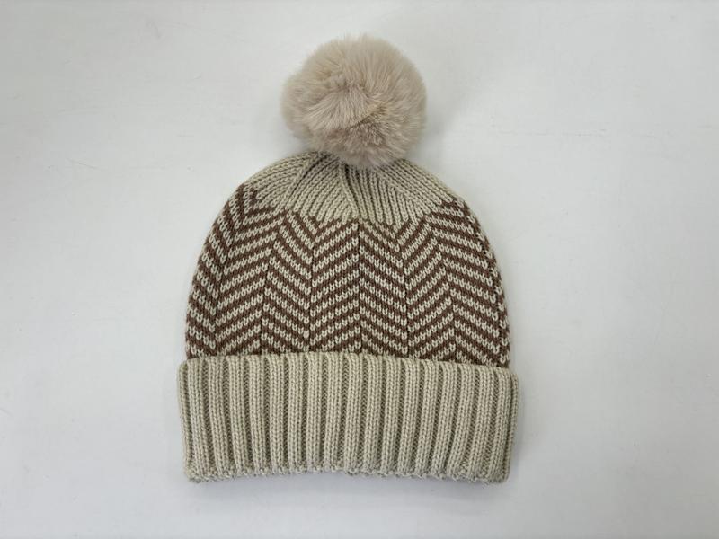 Crème gebreide muts met bruine chevron zigzagpatroon en pluizige nep-bontpompon op witte achtergrond.