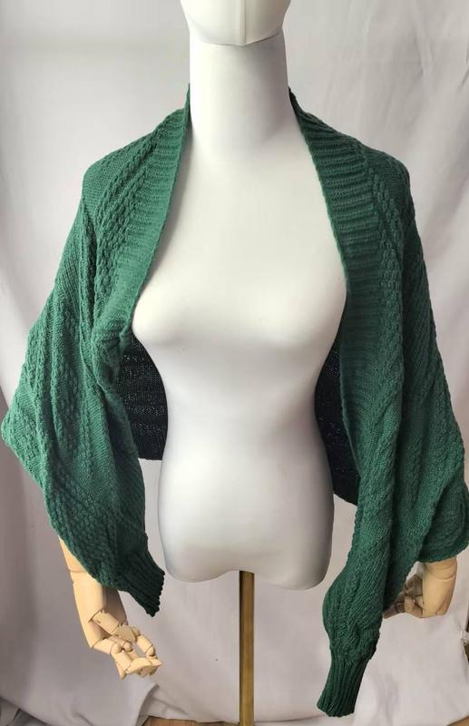 Groene kabelgebreide open cardigan shrug met ballonmouwen op een paspop.