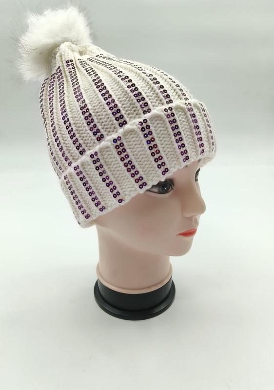 Witte geribbelde gebreide wintermuts met verticale paarse paillettenstrepen en pluizige neppels pompom.