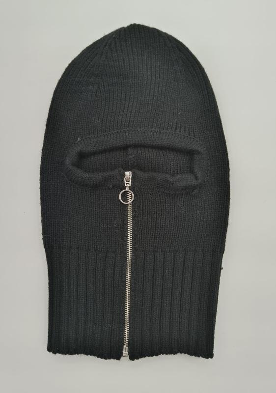 Zwarte geribbelde gebreide balaclava plat gelegd met zilveren rits voor gezichtsopening op grijze achtergrond.