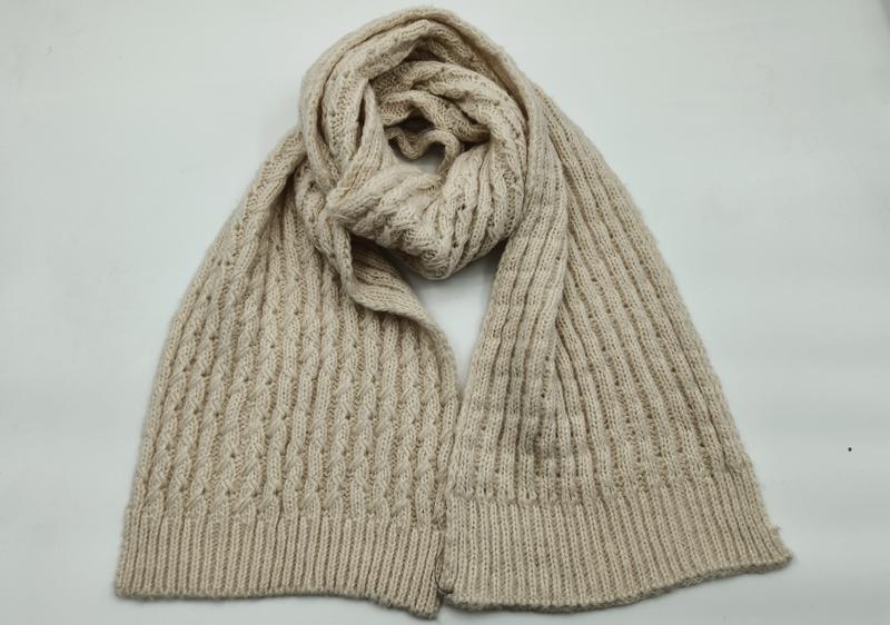 Crème beige kabelgebreide wollen sjaal losjes opgerold op witte achtergrond met geribbelde uiteinden.