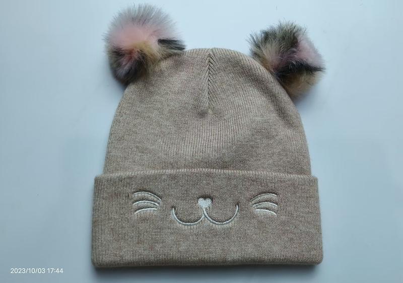 Beige gebreide kindermuts met twee multicolor pompons en geborduurd katgezicht