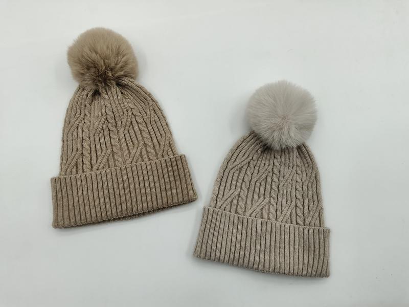 Twee kabelgebreide wintermutsen met pompons van imitatiebont in taupe en lichtgrijs op witte achtergrond.