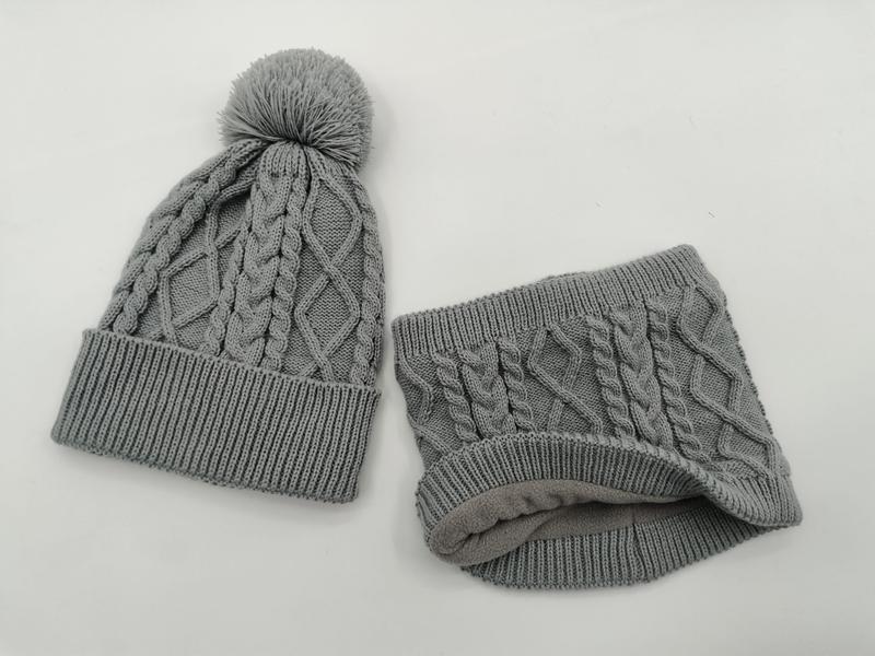 Grijze kabelgebreide wintermuts met pompon en bijpassende fleece nekwarmer set op witte achtergrond.