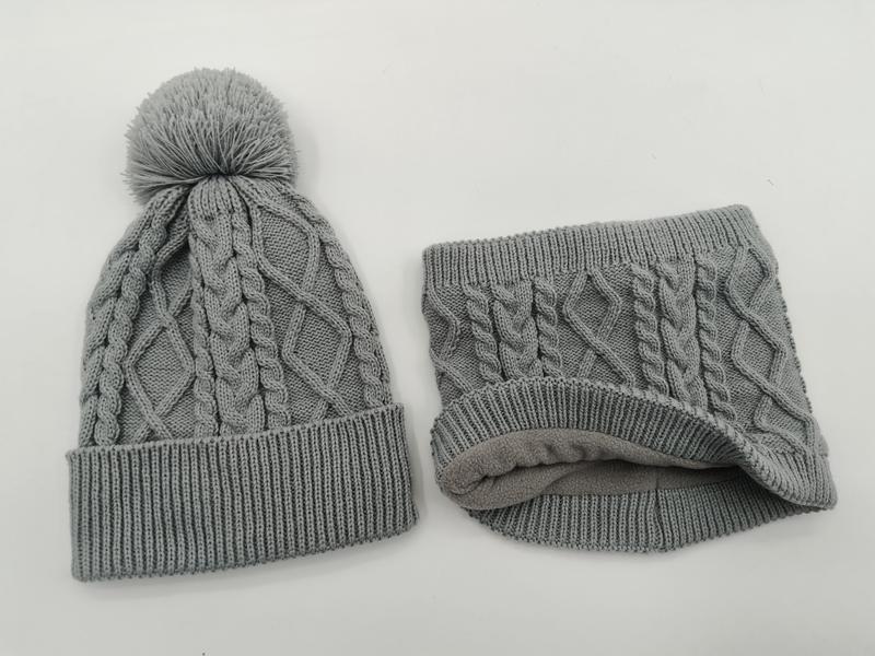 Grijze kabelgebreide wintermuts met pompon en bijpassende fleece nekwarmer set op witte achtergrond.