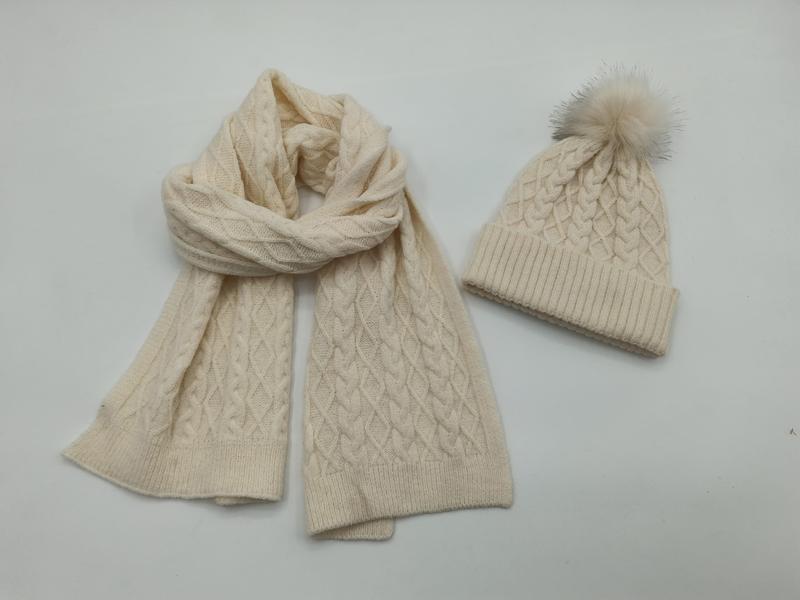 Crème gebreide wintersjaal en bijpassende muts met pompom op witte achtergrond.