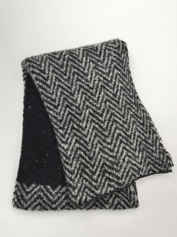 Gevouwen zwart-grijze chevron zigzagpatroon gebreide wollen sjaal met pluizige textuur op witte achtergrond.