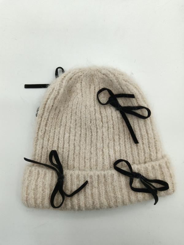 Crème gebreide wintermuts met grove ribstructuur en zwarte fluwelen strikken op witte achtergrond.