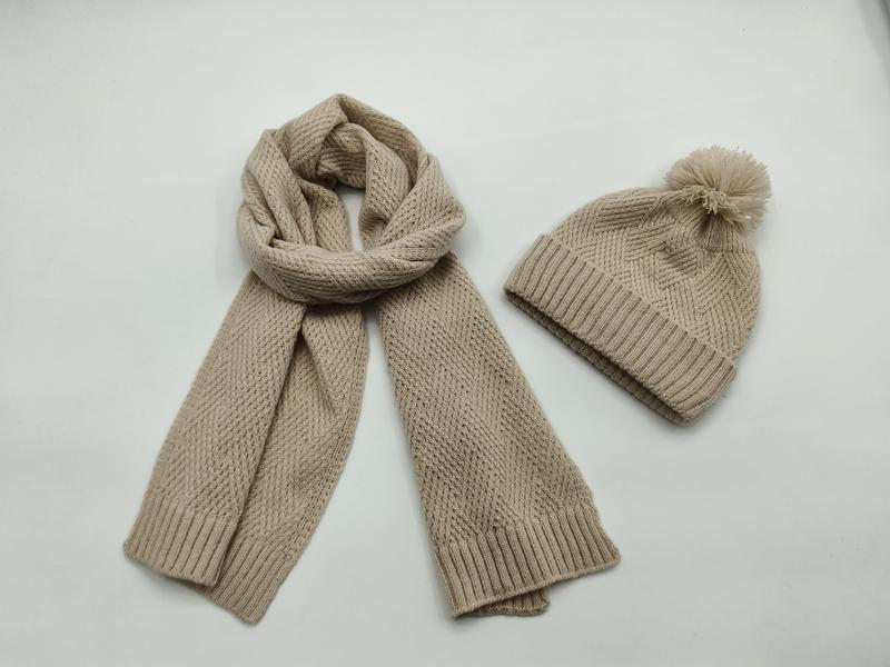 Beige grofgebreide sjaal en bijpassende muts met pompon op witte achtergrond.