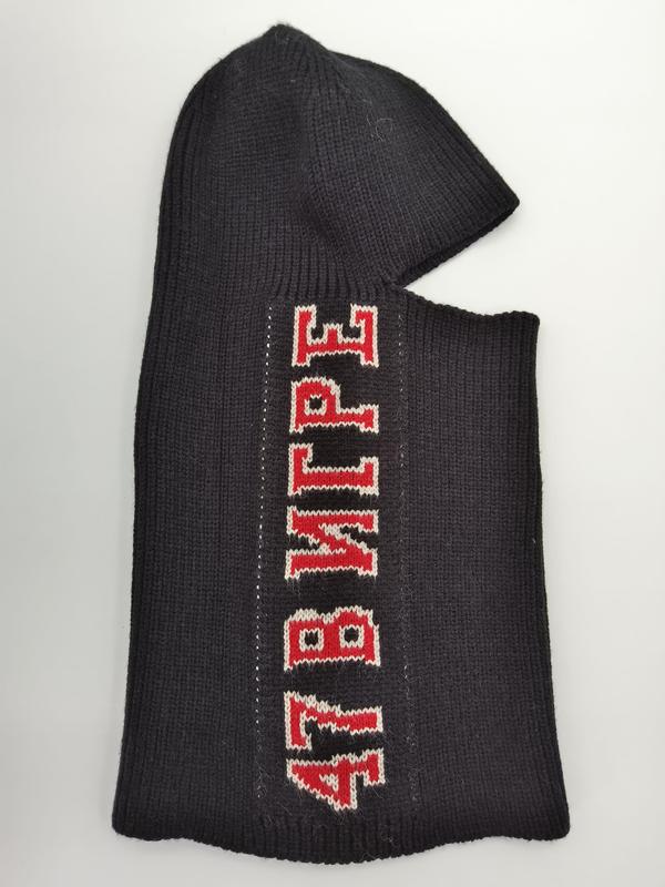 Zwart gebreide bivakmuts met rood en wit varsity-letterpatroon op donker wollen weefsel