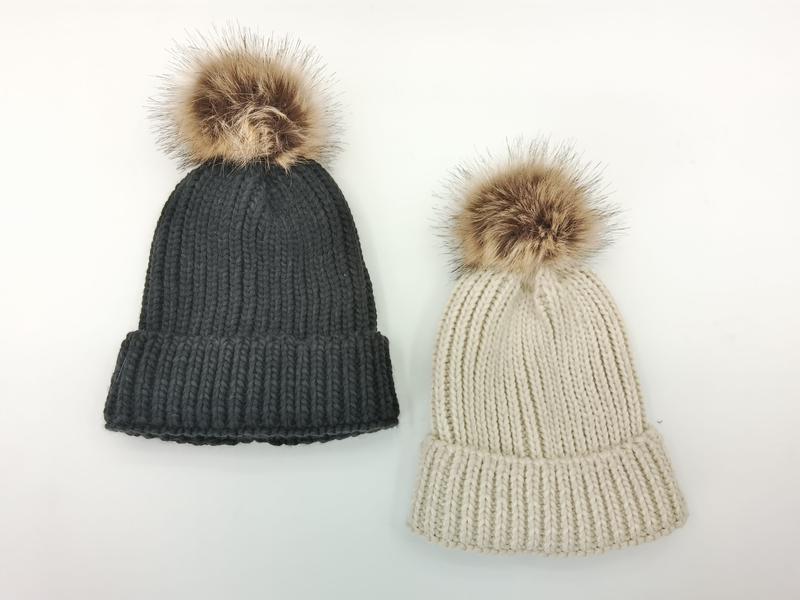 Twee grove gebreide mutsen met nepbont pompon in antraciet en beige op witte achtergrond.