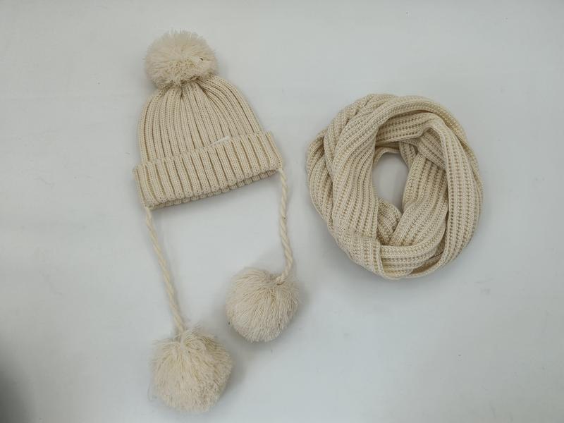 Crème geribde gebreide wintermuts met pompon en gevlochten banden naast bijpassende infinity sjaal.