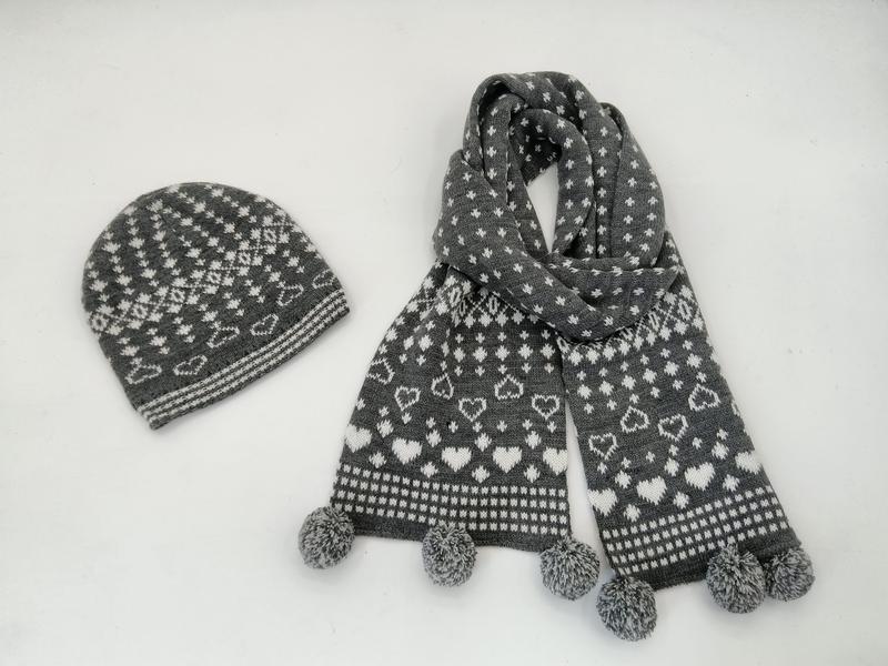 Grijze en witte gebreide muts en sjaal set met Fair Isle hartjespatroon en pompons op witte achtergrond.