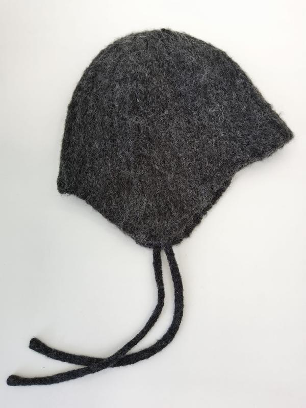Handgebreide houtskoolgrijs gevilte wollen babymuts met oorflappen en gevlochten stropdassen op witte achtergrond.
