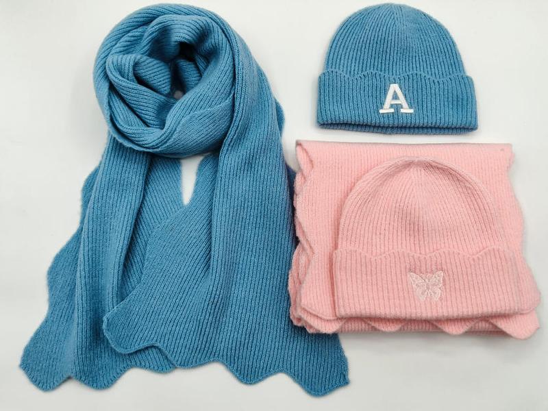 Blauwe gebreide sjaal met letter A-muts en roze vlinder-muts met sjaal set op witte achtergrond.