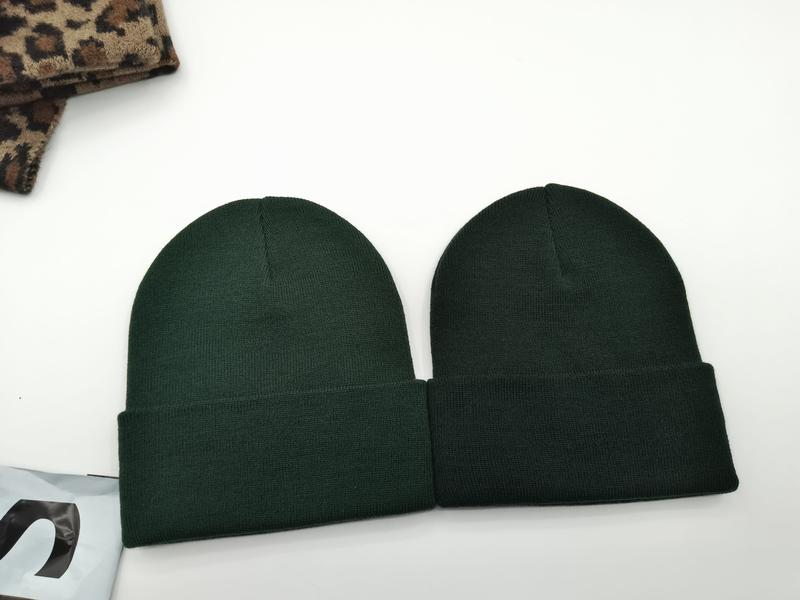 Twee donkergroene geribbelde gebreide beanie mutsen plat neergelegd naast luipaardprint stof op witte ondergrond.