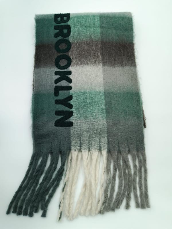 Gevouwen groen-grijs geruit wollen sjaal met ""Brooklyn"" borduursel en franjes op witte achtergrond.