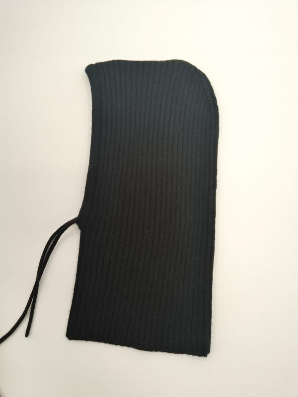 Zwarte geribbelde gebreide balaclava skimasker plat weergegeven op witte achtergrond met gebogen capuchonopening en trekkoorddetail.