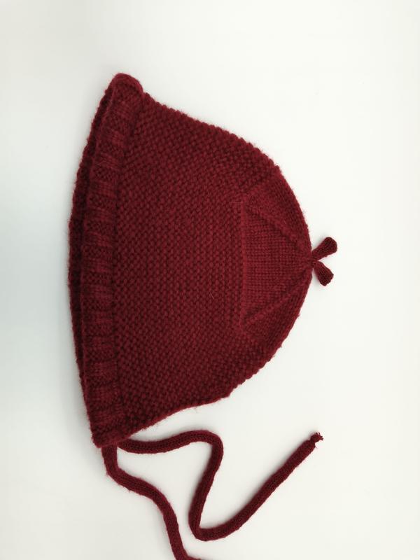 Handgebreid donkerrood babymutje met ruche rand, kinbandjes en puntige top, plat neergelegd op witte achtergrond.