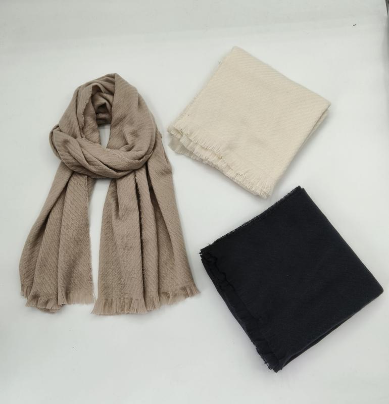 Drie sjaals op witte ondergrond: gedrapeerde taupe visgraatsjaal met franje, gevouwen crèmekleurige sjaal en gevouwen zwarte sjaal.