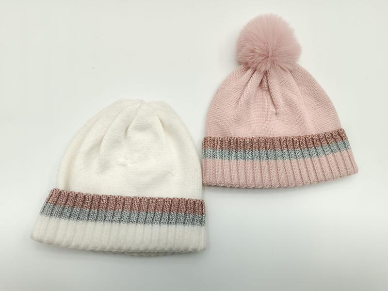 Twee gebreide mutsen in wit en roze met metallic streeprand, roze muts heeft een pluizige pompon bovenop.